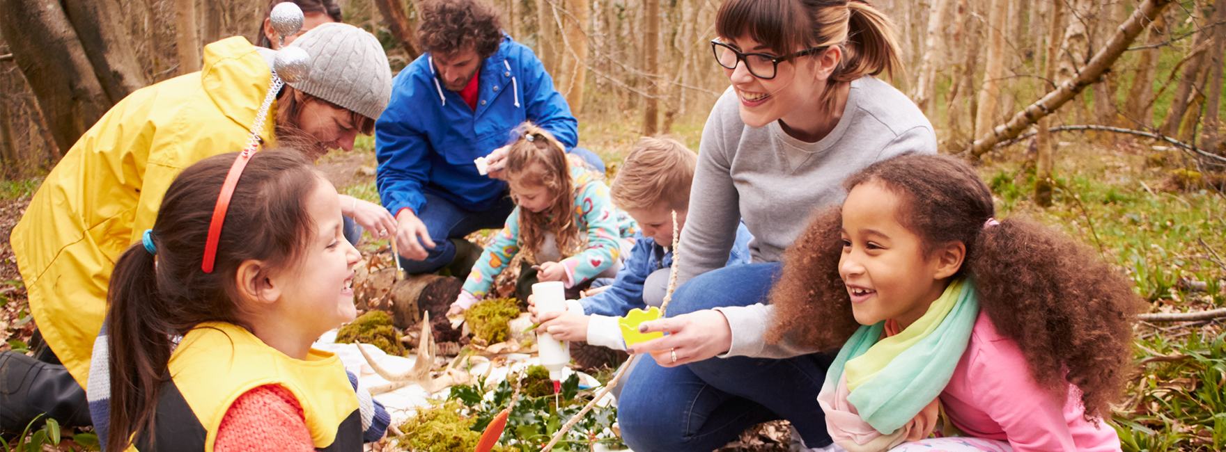 Kinder sitzen mit jungen Erwachsenen im Herbst auf dem Waldboden und erkunden Pflanzen und Pilze.