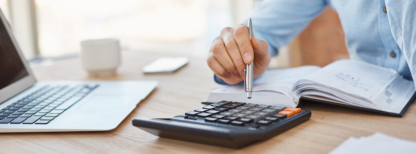 Blick auf einen Tisch, an dem eine Person sitzt und auf dem ein Notebook, eine Tasse, ein Kalender und ein Taschenrechner liegen.