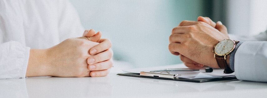 An einem Tisch sitzen sich zwei Personen gegenüber, zu sehen sind lediglich ihre Hände, die jeweils auf dem Tisch abgelegt sind.