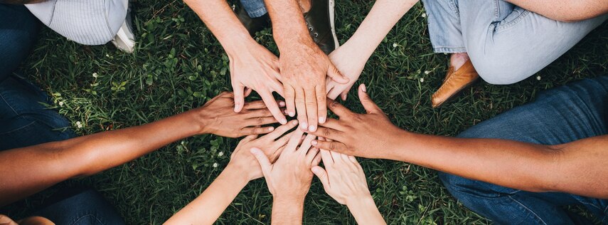 Aus der Vogelperspektive: Mehrere Personen sitzen auf einer Wiese im Kreis und halten ihre Hände in die Mitte.