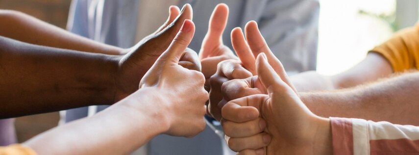 Personen stehen in einem Kreis und halten ihre Hände mit den Daumen nach oben in die Mitte.