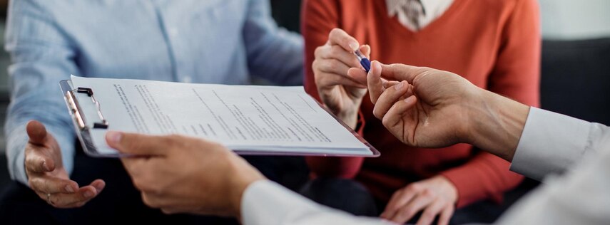 Zwei Personen wird ein Stift und ein Klemmbrett mit mehreren Zetteln überreicht.