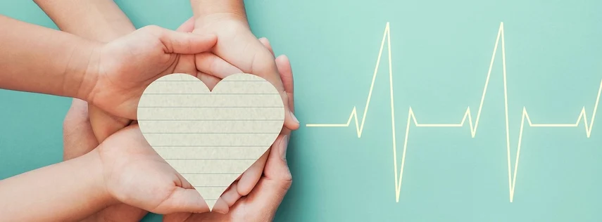 Mehrere Hände halten gemeinsam ein Herz aus Papier, rechts davon ist eine EKG-Kurve zu sehen.