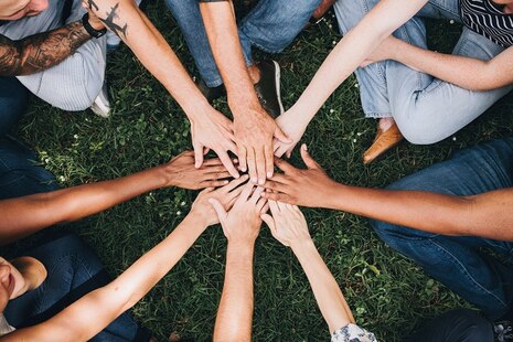 Mehrere Personen sitzen auf einer Wiese im Kreis und halten ihre Hände in die Mitte.