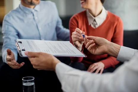 Zwei Personen wird ein Stift und ein Klemmbrett mit mehreren Zetteln überreicht.