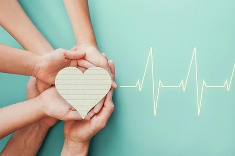Mehrere Hände halten gemeinsam ein Herz aus Papier, rechts davon ist eine EKG-Kurve zu sehen.
