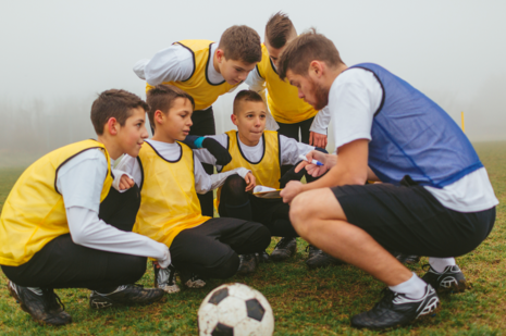 Ein junger Fußballtrainer hockt mit seinen Fußballern auf dem Rasen und bespricht die Taktik. Die Fußballer tragen gelbe Trainingshemdchen.